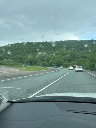 The image shows a view from inside a vehicle looking out at a highway. It is a cloudy day, and there are water droplets on the windshield. The highway is wet, indicating it may have rained recently or is currently raining. The road curves to the right, with several cars driving ahead. On either side of the highway, there is dense green vegetation and trees, forming a hilly landscape. There are also road signs indicating curves. The specific location in Usakly, Russia, cannot be determined from the visual information provided.