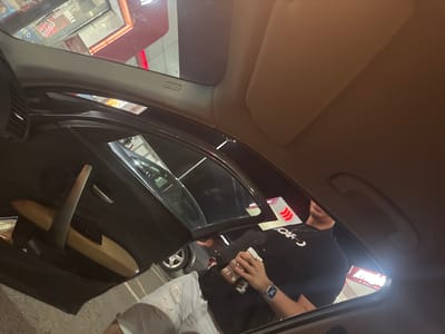 The image depicts the interior of a car, viewed from the driver's perspective. Outside the car, an individual wearing a black shirt and a watch is holding a cup. The car appears to be at a drive-through, where a box or container with red and white stripes is visible. In the reflection in the rearview mirror, a building with a red sign can be seen. Given the location constraints, there is no information to verify if the scene occurred in Vodenichane, Bulgaria.