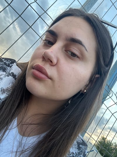 In Palanca, Moldova, a young woman with dark brown hair is captured in a close-up selfie. She is wearing a white shirt with a black and white patterned detail. Her hair falls around her face and shoulders. She has visible eyebrows, eyelids, and lips. A gold stud earring is visible in her right ear. The background consists of a metal fence with a grid pattern, behind which the sky is visible with clouds. The lighting suggests it is daytime, possibly late afternoon or early evening.
