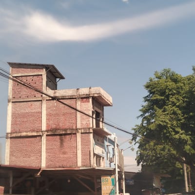 The image captures a daytime scene in Manetunong, Indonesia, featuring a multi-story building alongside a large tree against a clear sky. The building, constructed primarily with red bricks and concrete support beams, has an unfinished or industrial aesthetic, with visible rebar and exposed structural elements. Several black utility cables are strung across the front of the building.
To the right, a large, verdant tree with dense green foliage fills a significant portion of the frame, contrasting with the brick structure. The sky is bright blue with a single, elongated white cloud stretching horizontally across the upper part of the image, indicating sunny weather during the day.
At the ground level of the building, there are visible wooden supports and what appears to be a commercial frontage. A prominent green and yellow sign is visible on the lower right of the building, clearly displaying "BSI", "BANK SYARIAH INDONESIA", and "ATM". Below this, "AGUS NAWAR" is partially legible, likely a street or location name. Other unreadable signs are also attached to the building's facade. No people are visible, and no specific activities are taking place, presenting a static street scene.