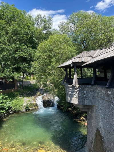 The image depicts a tranquil natural scene in Dulovine, Montenegro, likely captured on a clear, sunny day. Dominating the frame is a wooden structure with a weathered, shingled roof, supported by dark wooden beams and a rough stone wall. This structure appears to be a covered walkway or pavilion overlooking a vibrant, turquoise-colored pool of water.
Adjacent to the pool, a small waterfall cascades down rocky terrain, adding to the serene atmosphere. Lush green trees with dense foliage surround the water and the man-made structure, creating a sense of seclusion and natural beauty. The water is so clear that the rocky bottom is visible in some areas.
Below the wooden structure, there are glimpses of dark wooden benches, suggesting it's a place for visitors to rest and enjoy the scenery. A paved path or steps lead down towards the water. The overall impression is one of a peaceful, picturesque spot in nature, possibly a tourist attraction or a hidden gem within the Montenegrin landscape. There is no visible text in the image. The lighting suggests it is daytime, with bright sunlight illuminating the scene and casting shadows.