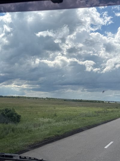 The image depicts a vast, open landscape under a dramatic sky filled with large, fluffy clouds. A paved road with a white line marking runs diagonally from the lower right corner towards the middle of the frame. To the left of the road, a field of green grass and wildflowers stretches into the distance, with a dense green bush on the far left. In the background, a tree line borders the field, beyond which are glimpses of distant buildings. The overall impression is one of a rural or semi-rural setting in the Астрахан ауылдық округі, Kazakhstan, possibly captured during a drive. The lighting suggests it is daytime, with areas of bright sunlight breaking through the clouds, casting varying shades on the landscape. There are no people visible in the image.