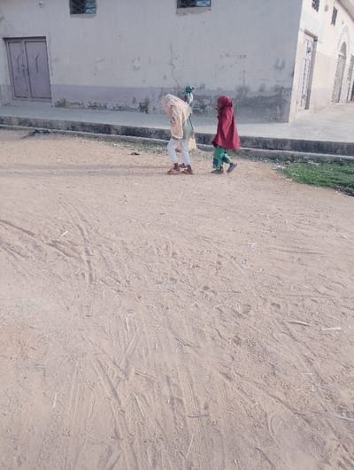 Two individuals, likely children, walk along a dirt road beside a light-colored building. The person on the left is mostly covered by a light tan shawl, while the person on the right wears a red headscarf and a knee-length red coat. The road shows signs of vehicular traffic, with tire tracks visible in the dirt. The building has a light-colored facade with a few windows and doors. A small patch of green vegetation is visible to the right of the road.
