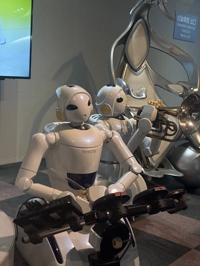 This is a medium shot photograph of two white robots, one slightly taller than the other, seated on what appears to be a display platform with a vehicle-like structure. The taller robot is on the left and has the word "TOYOTA" printed on its chest. This robot is posed as if controlling a vehicle with handlebars. The robot on the right, slightly behind and to the right, is playing a brass instrument, possibly a trumpet. Both robots have large, expressive eyes and circular yellow accents on the sides of their heads. The background is a gray wall with a sign in the upper right corner that reads "自動車館出口" and "Automobile Pavilion Exit" in English, along with other characters. The environment suggests an exhibition or museum setting, possibly at the Toyota Automobile Museum in Nagakute, Aichi Prefecture, Japan, which is near Nagoya. The lighting is artificial, and the overall scene is clean and well-lit.