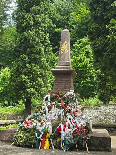 The image displays a memorial monument located in a lush, green outdoor environment in Ribaritsa, Bulgaria. The central object is a stone obelisk standing on a rectangular pedestal, constructed from a dark gray or brown stone. A golden-colored emblem is affixed to the obelisk section, depicting a Latin cross positioned above an open book, with a coiled serpent or scroll element beneath. The pedestal features an inscription in Cyrillic script; legible portions include "ГЕОРГИ БЕНКОВСКИ" (Georgi Benkovski) and the dates "1876" and "1908".

The base of the monument is heavily covered by a substantial collection of floral wreaths and bouquets. These tributes exhibit a primary color palette of red, white, and green, with additional accents of pink, purple, and yellow. Numerous white ribbons bearing Cyrillic text, some incorporating the Bulgarian national colors (white, green, red), are tied to the wreaths. Several of the larger wreaths are propped upright by thin wooden stakes.

The monument is situated amidst dense vegetation. Tall, columnar evergreen trees, consistent with conifers such as Thuja or Cypress, flank the monument on both sides. Various other deciduous trees form a thick, vibrant green backdrop. The ground directly surrounding the monument is paved with flat, irregular stone slabs. To the left, large, roughly hewn stone blocks form a low, linear structure, possibly a bench or retaining wall. To the right, a low wall constructed from dry-stacked, unmortared fieldstones is visible. The scene is illuminated by natural, diffused daylight, and no individuals are present in the image.