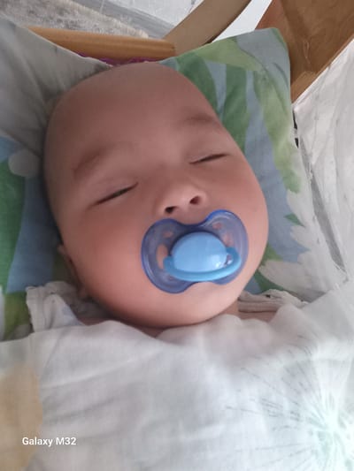 The media file is a close-up, top-down shot of a baby sleeping with a blue pacifier in its mouth. The baby is nestled in white blankets and lying on a colorful patterned surface, possibly a bedsheet or blanket, with stripes of blue, green, and yellow. The baby's eyes are closed, and its face appears peaceful.

In the upper left corner of the image, there is white text that reads "Galaxy M32," indicating the device used to capture the photo. The background is blurred, but suggests an indoor setting. The lighting appears to be natural, possibly from a window, and the overall mood is tranquil and intimate, capturing a moment of rest for the infant. The provided location context is "Ынтымак, Kyrgyzstan."