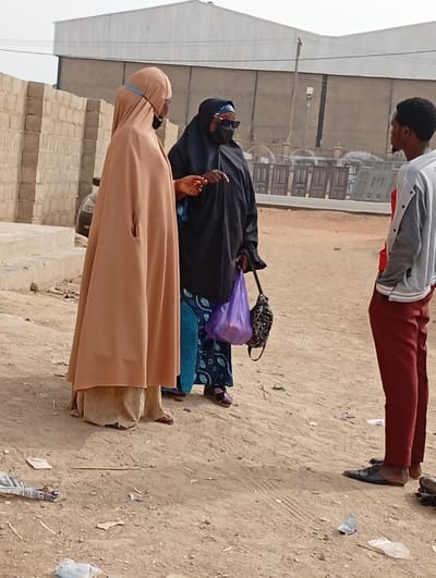 Three people are standing in a dirt lot near a building. Two women wearing face masks and modest clothing are facing a man. The woman on the left is wearing a tan head covering and the other woman is wearing a black head covering and sunglasses. The man is wearing a gray and white sweater and red pants. There are some building supplies and other items in the background.