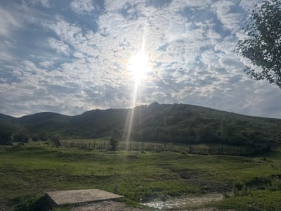 A sunburst penetrates a cloudy sky over a grassy hillside in Kashka-Suu, Kyrgyzstan. The sun, a bright white orb, is positioned slightly left of the center in the upper portion of the frame, emitting strong rays that create a dramatic lighting effect. Below the sky, a verdant hillside slopes upwards, covered in lush green grass and scattered trees. A weathered wooden fence, composed of vertical posts, runs horizontally across the middle ground of the image, delineating the terrain. In the foreground, a patch of green grass leads to a shallow body of water with reflections of the sky. A rough, rectangular concrete structure is visible in the lower left corner. The overall scene depicts a natural, rural landscape under a dynamic sky.