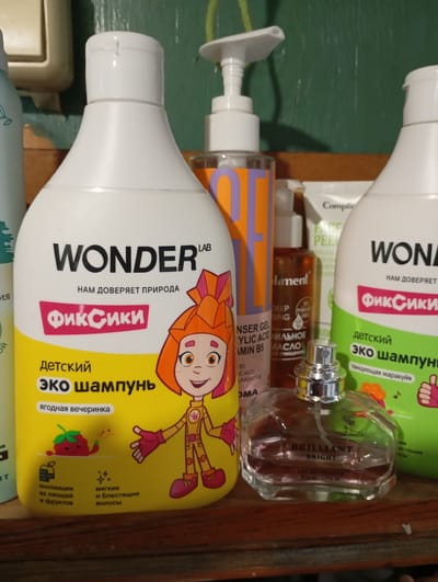 A close-up, eye-level shot captures a shelf adorned with various personal care products. In the foreground, a clear glass perfume bottle with a silver cap and a pinkish liquid is prominently displayed, its label reading "BRILLIANT BRIGHT EAU DE TOILETTE 50ML - 1.7 FL OZ". To the left, a large white shampoo bottle for children, branded "WONDER LAB", features a cartoon character and the text "ФИК-СИКИ" (Fixiki) and "детский эко шампунь" (children's eco shampoo), with additional text indicating "ягодная вечеринка" (berry party) and claims of "инновации из овощей и фруктов" (innovations from vegetables and fruits) and "мягкие и блестящие волосы" (soft and shiny hair). To the right of the shampoo bottle, another white bottle, partially obscured, carries the "WONDER" branding. Further to the left, a partially visible blue bottle with green leaf imagery is present. In the background, other bottles, including one with orange and purple branding and a pump dispenser, are visible. The shelf itself appears to be made of wood, set against a green, textured wall. The overall impression is of a home bathroom or storage area.