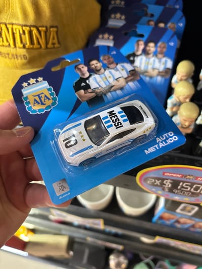 The image shows a person's hand holding a blister pack containing a white toy car with blue stripes. The car has the name "MESSI" printed on it in large black letters, and the number "10" with three stars above it. The packaging is blue and features the logo of the Argentine Football Association (AFA). Text on the packaging reads "AUTO METÁLICO" and "ARGENTINA". To the left of the car, there are cards with images of Argentine football players, also wearing the national team's jersey. In the background, blurred elements suggest a retail environment, possibly a store within the Aeropuerto Internacional Ezeiza, Argentina, with other merchandise and price tags visible. One price tag shows "$150" and "$95.00". The lighting suggests it's daytime, and the overall impression is of a souvenir or merchandise display in an airport.