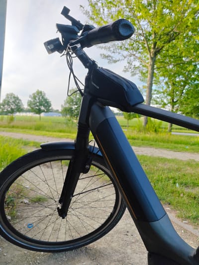 This close-up, low-angle shot features the handlebars and front wheel of a dark-colored electric bicycle parked outdoors. The bicycle's frame dominates the foreground, with its matte black finish and an orange accent strip visible on the down tube. A small black saddlebag, labeled "NEWBOLER," is attached to the handlebars. Two bicycle lights are mounted on the handlebars, one on each side. The front wheel, with its black rim and spokes, is prominent in the lower left of the frame.
The background suggests a natural, outdoor setting. Green grass and trees line a dirt path, hinting at a park or rural area. In the distance, more trees are visible, along with what appears to be a field and possibly some distant buildings or structures. The sky is bright and hazy, indicating daylight, likely late afternoon or early evening due to the warm quality of the light. The overall mood is peaceful and serene, suggesting a moment of rest during an outdoor excursion. The location context is Nieuw-Vennep, Netherlands. There is no visible text other than the "NEWBOLER" label on the saddlebag.