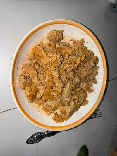 A close-up overhead shot shows a white bowl with an orange rim, filled with a textured, golden-brown food item that appears to be ground or finely chopped meat, possibly seasoned. The food is piled high in the center of the bowl. A fork with an ornate handle rests on the white surface below the bowl. A small, green object, potentially a piece of produce like a cucumber end, is visible at the bottom right of the image. The surface beneath the bowl and fork is a light-colored, tiled material. The image is sharp and well-lit, with the focus on the food and the table setting. No people are visible in the image. The location is Tuishevo, Russia.