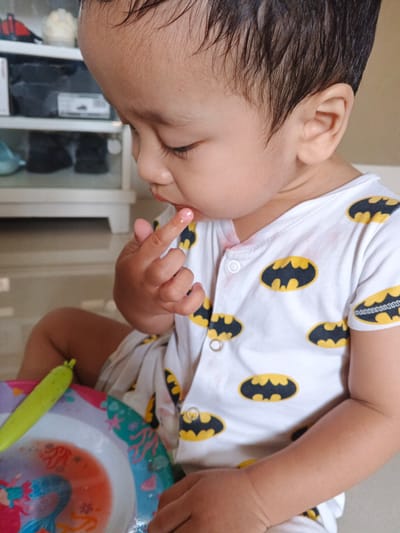 This media file features a close-up shot of a young child, likely a toddler, sitting down and engaged with a plate of food. The child is wearing a white onesie adorned with yellow and black Batman logos. The child's face is turned to the side, and they are bringing a finger, which appears to have a bit of pink food residue on it, towards their mouth.

In front of the child is a colorful plate decorated with mermaid and sea-themed graphics. A long, green object, possibly a piece of food like a green bean or a vegetable stick, rests on the plate. The background suggests an indoor setting with a blurred view of shelves containing various items, including what appear to be shoes. The lighting suggests daytime. The overall scene evokes a sense of a casual mealtime for a young child. No visible text is present in the image. The location context is Cibungbulang, Indonesia.