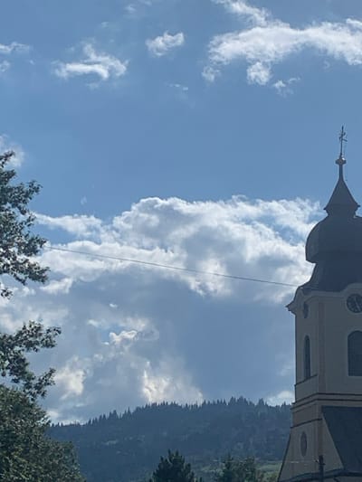 The image depicts a daytime scene in Yasinia, Ukraine, primarily focused on the upper portion of a church tower against a backdrop of mountains and a cloudy sky. The church tower, positioned on the right side of the frame, features a light-colored facade, a dark, domed roof, and a pointed spire capped with a cross. A circular clock face is partially visible on the tower. To the left, dark green foliage from a tree extends into the frame. A horizontal power line traverses the image, passing in front of a vast expanse of white and grey cumulus clouds. In the background, a dark, densely forested mountain range is visible beneath the cloud line. No individuals or discernible actions are present; the scene is static.