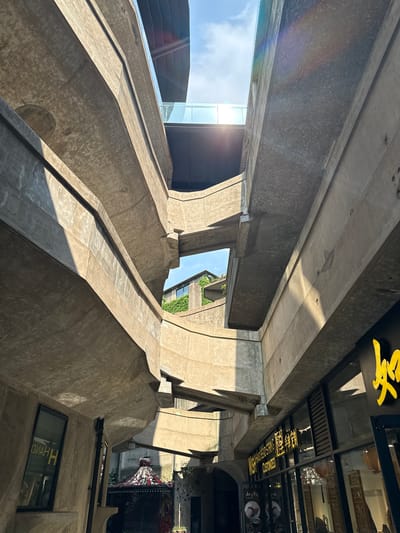 The image shows an upward view of a building structure with raw concrete elements in the Hongkou District of China. The building features prominent structural beams and pillars, some with a stepped design. Sunlight streams through openings in the structure, creating strong contrasts of light and shadow.
A modern building with glass elements is visible in the background. On the right, a building exterior is partially visible, showing signs in Chinese characters and decorative elements like flowers and lights. The windows on the building appear to have the word "HAND" displayed.
