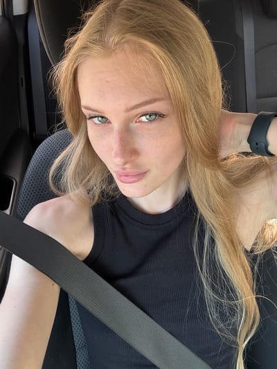 A young woman with blonde hair and green eyes is seated in the driver's seat of a car. She is wearing a black sleeveless shirt and a black seatbelt. The background shows the interior of a car, including a seat and a door. The woman is looking at the camera and has her right hand behind her neck. The lighting suggests it is daytime. There is no specific information about the location in the city of Krasnyy Voskhod, Russia provided in the image.