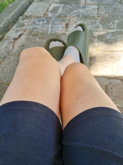 The image shows a person's legs from a high-angle perspective. The person is wearing dark blue shorts and light gray socks with dark green slide sandals. The legs are tanned and show some light leg hair. The sandals are simple and appear to be made of rubber. The background is a stone pathway, suggesting an outdoor setting. Sunlight casts shadows on the ground, indicating it is daytime. Some of the stones have faint chalk markings on them. The overall scene is casual and relaxed. The location is specified as Švenčionys, Lithuania.