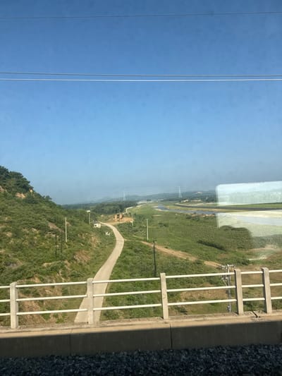 The image shows a rural landscape under a clear blue sky. In the foreground, there is a concrete barrier and a white railing, suggesting the vantage point is from a bridge or elevated structure. Below the railing, a paved road winds its way up a grassy, tree-covered hill on the left.
Further into the distance, the landscape opens up to a wide, flat area with a river or body of water meandering through it. Patches of green fields and some scattered trees are visible. In the mid-ground and background, there are several utility poles and what appear to be power lines stretching across the horizon. A tall transmission tower is also visible in the distance. The overall impression is of a serene, undeveloped area with some infrastructure. It is not possible to determine the exact location within Huludao City, China, from the image alone. There are no people or animals visible in the scene.