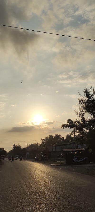 The image depicts a street scene at dusk or dawn in Sumbokrayek, Indonesia. The sun is low on the horizon, partially obscured by clouds, casting a warm, golden light across the asphalt road. The road appears wet, with numerous small reflections of light shimmering on its surface. A line of trees and some small buildings are visible along the right side of the road, with a darker silhouette of foliage dominating the upper right portion of the frame. Several motorcycles are present on the road; some are moving, indicated by blurred motion, while others appear parked. One motorcycle is visible parked under a small structure on the right. A single bird is captured in flight in the upper left quadrant of the sky. The sky itself is filled with a mix of light and darker clouds. A thin electrical wire stretches horizontally across the upper part of the image.