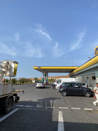 The image depicts a sunny day at a gas station in Boukadir, Algeria. The gas station has a bright yellow canopy with blue and yellow accents. Several cars are present, including a silver sedan, a dark gray hatchback, and a white van. A person is visible near the gray hatchback, bending down. Another person is walking away from the camera to the right. In the foreground, the back of a flatbed truck with a lift mechanism is visible. The ground is paved with interlocking bricks. The sky is blue with scattered white clouds. In the background, there are trees and shrubs, and a hillside. A sign with the logo "JD NAFTAL" is prominently displayed on the gas station canopy.