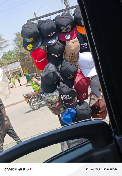 The image captures an outdoor street scene on a very bright, sunny day, viewed from inside a vehicle, likely through an open window. The primary subject is a dense display of baseball caps and trucker hats hanging from a makeshift metal rack, presumably for sale by a street vendor.
In the immediate foreground and framing the right side of the image, dark components of the vehicle's interior are visible, including a vertical pillar and a horizontal armrest or window sill. Below the hats, a reflection of a person wearing a plaid shirt (presumably the photographer) can be seen in the vehicle's window glass.
The hats, numbering over twenty, are diverse in color and brand. Many are black or dark-colored, interspersed with red, tan, white, blue, maroon, and camouflage patterns. Several prominent brand logos and designs are visible:
* Multiple hats bear the "FILA" logo.
* A black hat with a red brim features a white "23" in a jersey-style font.
* Another tan hat prominently displays the "MERCEDES-BENZ" logo.
* A red hat has "SPORTSCAP" written on it.
* A black hat with a golden logo depicts a basketball player, reminiscent of the Jumpman logo.
* A red hat has "Deluxe" written on it.
* A black hat features "LA," commonly associated with the Los Angeles Dodgers.
* Other hats display "TOYOTA" and "Panosonic Toyota Racing" logos.
* A camouflage hat has a patch resembling the American flag.
* Some hats are solid colors, while others are trucker-style with mesh backs.
Beyond the hat display, the background reveals a bustling street or roadside environment. The day is clearly bright and sunny, indicated by harsh shadows and strong illumination. A person wearing a bright light green or yellow reflective vest is seated on a motorcycle or scooter, facing away from the camera, on what appears to be an unpaved or worn asphalt road. Further into the background, other individuals, some also wearing reflective vests, are visible near simple buildings or structures. Trees and sparse vegetation line the distant parts of the scene, under a clear blue sky.
The overall impression is that of a street market or roadside stall, likely in a warm climate, with the caps being sold to passersby or from a stationary vehicle.
Visible text in the image includes the brand names and logos on the hats as described above. Additionally, technical information about the photograph is overlaid at the bottom: "CAMON 40 Pro" (indicating the device used) and "53mm f/1.8 1/828s ISO50" (camera settings).