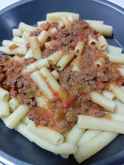 A close-up shot shows a serving of pasta with meat sauce, served in a dark grey square dish. The pasta is tube-shaped, likely rigatoni, and arranged in a somewhat haphazard pile with some noodles at the bottom and the rest layered on top. The sauce is a reddish-brown color and appears to contain ground meat, tomatoes, and possibly some chopped vegetables. The sauce is distributed unevenly, covering the top portion of the pasta. A silver fork is visible, slightly protruding from the pasta and sauce on the left side.
This image was not taken in Yukariyuz, Uzbekistan, as it does not contain any information that would indicate location.