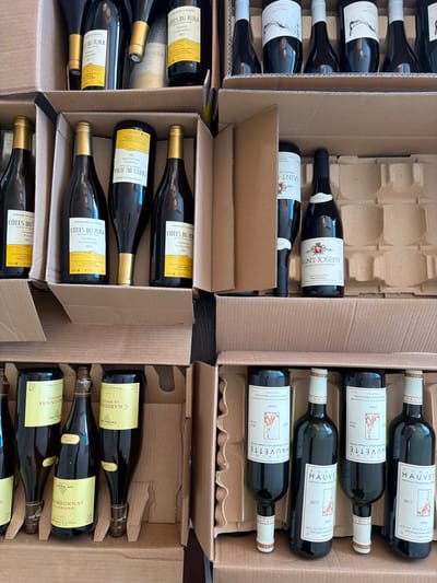 The scene, located in Marche-lez-Écaussinnes, Belgium, depicts an overhead view of multiple open corrugated cardboard boxes filled with wine bottles. Approximately six sections of these boxes are visible, containing numerous dark-colored glass bottles. Identifiable labels include "COTES DU JURA" (yellow labels, showing vintages 2022 and 2023), "SAINT-JOSEPH 2023" (white label featuring a red and white crest), "CHARDONNAY" (light yellow labels, vintages 2022 and 2023), and "DOMAINE HAUVETTE DOIX 2017" (white labels with a stylized tree graphic). Other bottles display white labels with abstract black designs or have obscured labels. All bottles are systematically secured within the internal cardboard dividers of their respective boxes. The boxes are arranged on a dark, flat surface. No individuals or external interactions are observed.