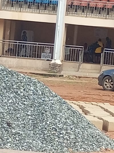This image depicts an outdoor scene in Ajalli, Nigeria, alongside a two-story building. In the immediate foreground, to the left, is a substantial pile of light grey aggregate (gravel). To its right, on a reddish-brown dirt surface, lie multiple rows of light-colored rectangular blocks, possibly concrete pavers. The front wheel and lower body of a dark-colored vehicle with a multi-spoke silver rim are partially visible on the far right, parked on the dirt.
The midground features the ground level of a light-colored building, presenting an open veranda or walkway shielded by a silver metal railing. On the left side of this veranda, a single individual dressed in dark clothing stands near a large, light-colored rectangular object. Further to the right within the veranda, a group of at least three individuals is present. One person wears dark attire with a distinct yellow garment or pattern over it, while others are in darker clothing. Their specific actions are indistinct. A small white sign with dark script is affixed to the metal railing in the center of the veranda. A tall, light-colored pole, featuring a stone-clad base, rises vertically from the ground in front of the building. The upper level of the building displays rows of empty stadium-style seating, with visible red and blue seats, behind a metal railing. The ground throughout the scene is reddish-brown earth.