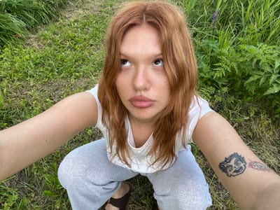 A young woman with long, reddish-brown hair and fair skin is crouching on a grassy area, looking up and to her left. She is wearing a white tank top and light gray patterned pants. A tattoo of a cartoonish face is visible on her right arm. The background is filled with lush green grass and foliage, suggesting an outdoor, possibly rural or park-like setting. There are no identifiable landmarks or structures that would indicate a specific location in Суроватиха, Russia. The lighting suggests it is daytime, likely under natural sunlight.
