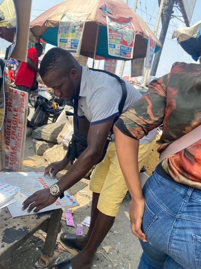 The image depicts an outdoor market scene in Aiyetoto-Asogun, Nigeria, under bright daylight. In the foreground, an adult male with short dark hair, wearing a light-colored short-sleeved polo shirt, yellow shorts, and black slides, leans forward, his attention focused downwards. He manipulates a clear plastic bag containing small items, which rests on a sheet of cardboard displaying repeated "FRAGILE" text in red, placed on a rudimentary wooden table or bench. A silver-toned watch is visible on his left wrist. To his right, partially obscured, stands another adult, wearing a multi-colored patterned long-sleeved shirt and blue denim jeans. A light-colored strap crosses their shoulder.

In the background, a large, multi-paneled market umbrella, colored brown and light blue/white, provides shade. Several printed banners are attached to the umbrella, advertising financial services such as "CASH WITHDRAWAL," "CASH TRANSFER," and "UTILITY BILL" alongside various bank logos. Further to the left background, a person in a red shirt is partially visible near what appears to be a bicycle or motorcycle. The ground is uneven, dusty, and littered with rocks, cardboard pieces, and small colorful wrappers. A dark bag and piles of rubble are visible behind the central male.