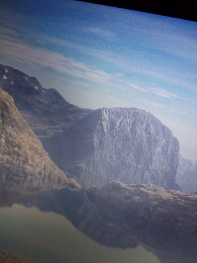 The image is a photograph of a mountain range with a body of water in the foreground, taken from an elevated perspective. The sky is visible in the upper portion of the image, with some wispy clouds. The mountains are rocky and appear to be covered in patches of vegetation. The water in the foreground is dark and reflects the surrounding landscape. The image quality is somewhat grainy, suggesting it may have been taken with a low-resolution camera or under low-light conditions. The image does not depict the city of Mbar, Nigeria, or any urban elements.