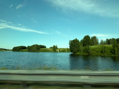 The image shows a wide, still body of water, likely a lake, with a shoreline lined by dense green trees and rolling hills. The sky is a clear, bright blue with scattered white, wispy clouds. In the distance, a small building with a light-colored roof is visible nestled amongst the trees on the far bank. The foreground of the image is dominated by a blurred, horizontal metallic barrier, suggesting the photo was taken from a moving vehicle, possibly a car or train, traveling alongside the water. The reflections on the water's surface indicate it is calm with gentle ripples. The overall scene depicts a serene, natural landscape under a bright sky. The location is identified as Posevnaya, Russia.