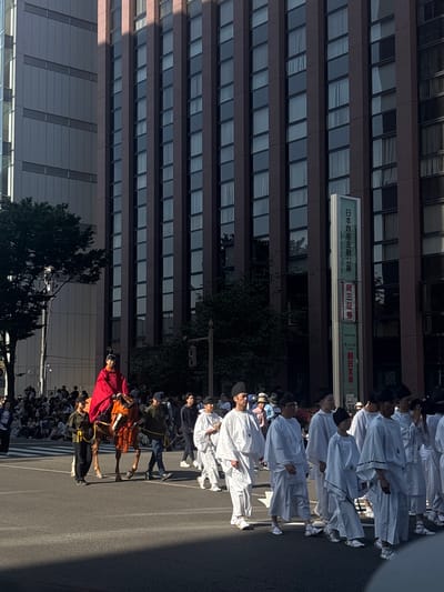 A daytime parade or procession unfolds on a paved city street in Kanazawa, Japan. The precise location is identifiable by a tall, vertical building sign on the right displaying "日本政策金融公庫" (Japan Finance Corporation), "岡三証券" (Okasan Securities), and "朝日生命" (Asahi Life), placing the scene likely on Hirosaka Dori near Korinbo.
A central figure, adorned in a prominent red cloak and dark traditional headwear, rides a reddish-brown animal, possibly a horse, decorated with orange and red fabric. An individual in dark attire and a cap walks alongside the animal's left, appearing to guide it. In the right foreground, a group of approximately eight individuals dressed in matching white traditional Japanese garments, dark headwear, and white traditional sandals walks in a disciplined formation from right to left. Other procession participants are visible behind the animal. On the far left, numerous spectators, some seated, observe the event from the side of the street.
The urban environment includes a prominent multi-story building on the right with a dark brown facade and numerous regularly spaced rectangular windows, reflecting elements of the sky. To the left, behind a large green tree, another tall building with a lighter grey, panelled facade is visible. Long, distinct shadows extend across the street from right to left, indicating a low sun angle, suggesting either morning or late afternoon. A traffic light pole is partially visible on the right.