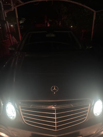 A dark Mercedes-Benz car is parked at night in Kableshkovo, Bulgaria, possibly under a carport or overhang structure. The car's headlights are on, illuminating the front grille and hood. The background is dark and indistinct, with some blurry structures and foliage visible. There are no people present in the image.