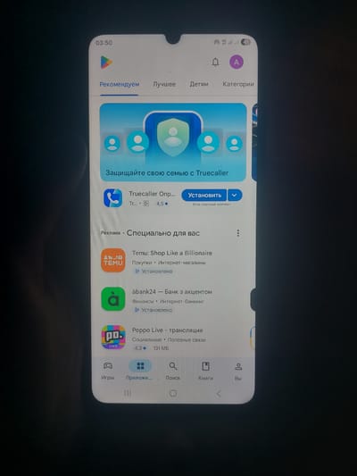 The image shows a close-up of a smartphone screen displaying the Google Play Store app. The phone is likely held by a person, as indicated by the finger partially visible on the left side of the screen. The app store features a banner at the top with the text "Защищайте свою семью с Truecaller" (Protect your family with Truecaller). Below this, there are various app listings including "Temu: Shop Like a Billionaire" and "abanku24 - Bank with an account," both marked as "Установлено" (Installed). The bottom navigation bar of the app store shows icons for "Игры" (Games), "Приложе..." (Apps...), "Поиск" (Search), "Книги" (Books), and "Вы" (You). The time displayed on the phone's status bar is 03:50, and signal indicators show "4G" and Wi-Fi. The background is dark, suggesting the photo was taken in low light conditions. There is no information in the image to suggest the location is the city of Korolevo, Ukraine.