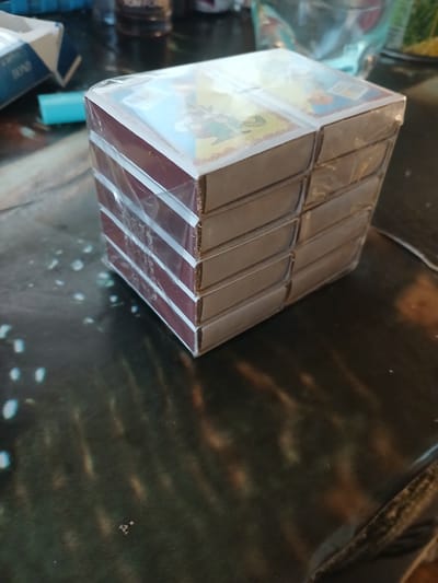A stack of six card decks, shrink-wrapped together, sits on a dark, reflective surface. The decks appear to be for playing cards, with the top decks showing portions of colorful illustrations featuring cartoon characters. The edges of the decks have a burgundy colored band with white stripes. The shrink wrap is slightly crinkled and reflects light, creating bright white streaks. In the background, out of focus, is a blue and white cigarette pack labeled "BOND." To the left of the card deck, a small, bright blue object, possibly a cigarette lighter, is visible. Further in the background, a blurry glass container and other unidentifiable objects are present. The scene is lit by artificial light, creating reflections and highlights on the surfaces. There are no people or animals present in the image. The location is Hlushky, Ukraine.