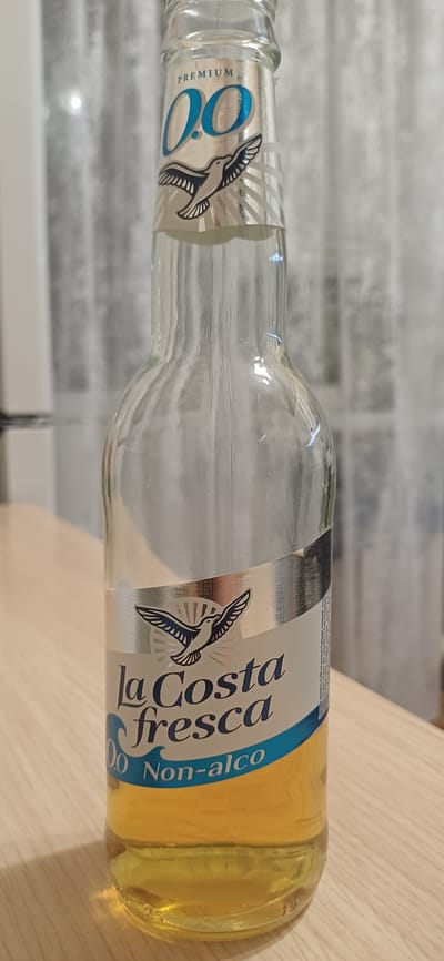 A clear glass bottle containing a light yellow liquid stands upright on a wooden surface. The bottle has a label with text and a graphic of a bird in flight. The background consists of sheer, light-colored curtains. The image is a close-up shot, focusing on the bottle. There is no information in the image to suggest it is located in Zuevka, Russia.