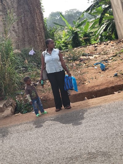 In Puerto López, Colombia, an outdoor scene captures an adult female and a young child standing on the dirt shoulder adjacent to an asphalt road. The woman, positioned centrally, is a dark-skinned individual wearing a light-colored, sleeveless collared shirt, dark trousers, and dark sandals. She carries a grey shoulder bag on her left side and holds a blue mesh bag containing several blue containers in her right hand. Her head is turned to her left, looking towards the background. To her left and slightly in front, a dark-skinned young boy, wearing a dark brown long-sleeved shirt, blue jeans, and light green open-toed shoes, stands holding a small dark object in his left hand while extending his right arm to the left. His head is slightly turned towards the woman.
The environment features a grey asphalt road occupying the foreground. Immediately behind the subjects, a dirt embankment rises, characterized by exposed earth and sparse vegetation, including tall grasses and broad-leafed plants. Various items of litter, including blue plastic bags and a purple cloth, are visible on this embankment. To the left, a tall, textured concrete wall or structure is heavily overgrown with vines and other vegetation. On the far right, another partial concrete structure is visible. The background consists of dense tropical-like foliage, including banana trees, under a bright sky. Shadows are cast on the asphalt road, indicating a daytime setting.