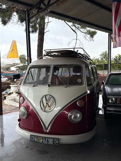 The image shows the front of a maroon and white Volkswagen Kombi van parked inside a building. The van has a roof rack and a South African license plate reading "HZT 276 EC." A portion of an American flag is visible in the upper left corner. In the background, a partial view of a yellow and white airplane tail fin and another black vehicle can be seen. The van is parked near a wall inside what appears to be a garage or storage area. Based on the license plate and the context of the provided information, the location is likely Kraaifontein, South Africa. There are no people visible in the immediate foreground, though there is a person visible through the front windshield of the bus.
