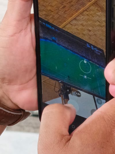 An overhead, close-up, vertical shot captures a person's hands holding a smartphone. The phone is oriented vertically, and its screen displays a live camera feed. The phone's screen is divided into two sections, with the top section showing a drone's perspective of a green field, possibly a soccer field, with blue markings and a yellow object in the center. A white circle with a "6" inside is visible on the field. The bottom section of the screen appears to be a camera interface, showing zoom options like "2.8x" and other icons. The background of the image consists of a woven bamboo ceiling and the hands of the person holding the phone, suggesting an indoor environment. The specific location in Julikeude Dua, Indonesia, is not discernible from the image itself.