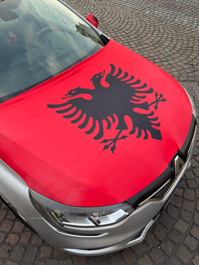 The image shows the hood of a silver car covered by a red cloth featuring a black double-headed eagle, the national symbol of Albania. The car is parked on a cobblestone street. In the background, through the windshield, a small Albanian flag can be seen hanging from the rearview mirror, and another is attached to the car's side mirror. The setting appears to be a public area in a city, with cobblestones suggesting an older urban environment. While not explicitly stated to be in Parma, Italy, the presence of Albanian national symbols on the car suggests a possible connection or event involving the Albanian community in that city. The car itself is modern, with visible headlights and grille. The overall impression is one of national pride or support being displayed.