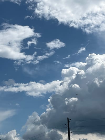 This is an outdoor, eye-level, vertical photograph of a blue sky with scattered clouds and a utility pole. The sky is a vibrant blue with fluffy white cumulus clouds of various sizes. Some clouds are wispy and thin, while others are large and dense, with shadowed undersides. In the lower right portion of the image, a dark wooden utility pole stands with several wires stretching horizontally across the frame. The pole has insulators attached to it, supporting the wires. The lower half of the image is darker due to the dense cloud cover and the presence of the utility pole and wires. There are no people visible in the frame. The location is Pamyati 13 Bortsov, Russia.