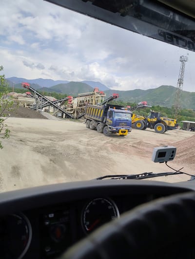 The image, taken from the interior of a truck, shows a construction or mining site in Cherniche, Bulgaria. Visible outside the windshield is a blue dump truck and a yellow Komatsu WA470 wheel loader. In the background are mountains, conveyor belts, and processing machinery. A tall communication tower is also visible. The ground is dirt and gravel. The truck from which the photo is taken has a dashboard with a speedometer and electronic display visible. A toll pass transponder is mounted on the windshield. The sky is cloudy.