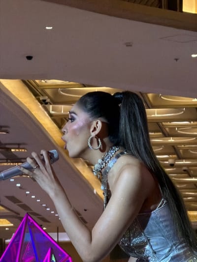 The media file shows a medium shot of a woman performing on a stage, with her head bowed and facing left. She has long, dark hair pulled back and is wearing a shimmering silver outfit adorned with rhinestones and a large, elaborate necklace. Her makeup is visible, with dramatic eye makeup and glitter on her face. She is holding a microphone in her right hand, which is resting on a railing. Her left hand is also visible, with a ring on one finger. The background is blurred, but appears to be an indoor venue with reflective surfaces and lighting that suggests a performance or concert. The overall mood seems to be one of intense focus or emotion during the performance. The specific location in Cebu City, Philippines is not explicitly evident in the image itself, but could be inferred from external context.