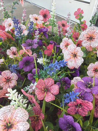 The image displays a close-up view of a vibrant floral arrangement, likely artificial due to the uniform texture and crisp detail of the petals. The arrangement is dense, with various flowers in shades of pink, purple, blue, and white, interspersed with green foliage. The pink flowers exhibit a delicate pattern of black lines on their petals, while the purple flowers are deep and velvety. Small clusters of blue and white flowers add texture and contrast. The background is blurred, suggesting a shallow depth of field, with hints of a light-colored, possibly building facade and a vertical line that could be a door or window frame. The lighting is natural and even, casting no harsh shadows. There are no people, animals, or discernible human activity in the image. The setting does not provide any specific clues about its location within the city of Lebanon, United States of America, or any urban environment.