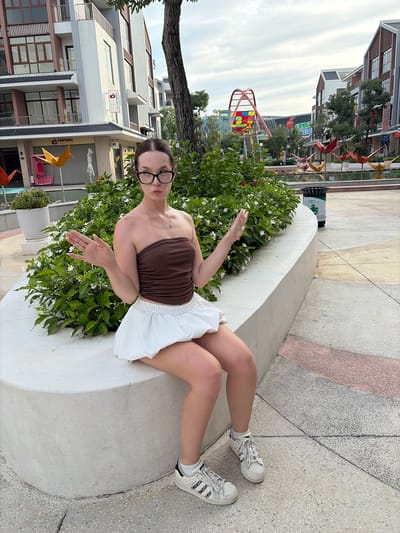 A young woman with dark hair pulled back and wearing glasses sits on a low, curved, concrete wall. She has on a brown strapless top and a white bubble skirt, and white sneakers with black stripes. Her arms are outstretched, palms up, in a questioning gesture. Behind her is a dense green bush with small white flowers and beyond that, buildings and an amusement park ride structure. The ground is paved with grey and pink tiles.