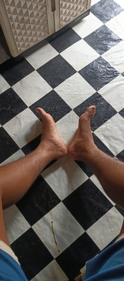 The image depicts a top-down view of a person's bare feet and lower legs against a black and white checkered floor. The floor appears to be linoleum or a similar material and shows signs of wear, with some white squares appearing slightly stained or textured. A beige appliance with a patterned surface and the word "TABITHA" visible on its side is present in the upper portion of the frame, casting a slight shadow on the floor. A small brown stain is visible on one of the white squares of the floor. The person's skin is visible on their feet and lower legs, which are hairy. The person is wearing blue fabric clothing around their lower body, partially visible in the bottom corners of the image. The scene appears to be indoors, possibly in a residential setting. The location is identified as Kemuning, Indonesia.