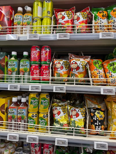 The image displays a shelf in a supermarket located in Sokilnyky, Ukraine, stocked with various food and beverage items. The shelves are filled with an assortment of products including bottled aloe vera drinks in green and brown hues, canned sparkling beverages in red and yellow packaging featuring Korean script, and bagged snacks. Some of the snack bags have red and white cartoon characters on them, while others have orange and yellow packaging with Chinese characters. Price tags are visible on some of the items. The lighting suggests an indoor commercial environment. There are no people visible in the frame.