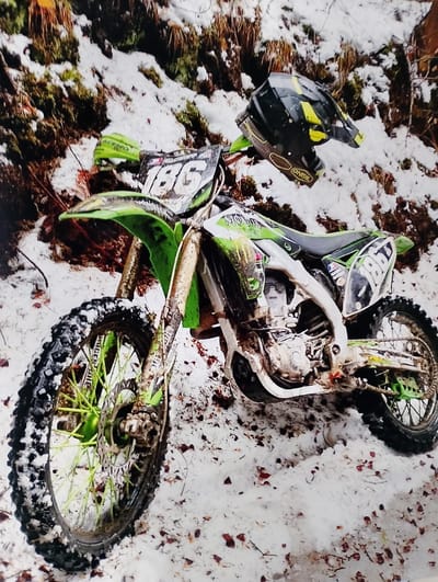 A green and white motocross dirt bike, heavily caked with mud and snow, is the central subject of this outdoor photograph. The bike displays the number "186" prominently on its front and side plates. A black and yellow motocross helmet, bearing the "ONFIL" (likely O'Neal) brand, is placed on the handlebars.
The setting is a snowy, natural environment, possibly a forest or a rugged trail in Ribnovo, Bulgaria. The ground is covered in a layer of white snow, through which patches of dark moss, brown earth, and scattered reddish-brown leaves and debris are visible. The background shows hints of tree roots and dense vegetation, typical of a wooded area. The dirty condition of the bike and its tires suggests it has been actively ridden through mud and snow, indicating an off-road adventure or race. The presence of the helmet on the handlebars implies the rider has recently dismounted or is about to embark on a ride. The lighting appears to be natural daylight, possibly under an overcast sky. Partial branding, including "MONSTER" (Monster Energy), can be seen on the bike's body.