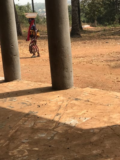 The image depicts an outdoor scene in Egboje, Nigeria, under clear daylight. In the midground, an adult female with dark skin walks from left to right across a reddish-brown dirt path. She is balancing a white, shallow basin on her head and wears a multicolored patterned garment, a dark top, a red headscarf, and light-colored flip-flops. A yellow satchel or bag is visible on her back. Blue markings are present on her face. The foreground is partially framed by two large, textured grey concrete pillars casting shadows onto a dirt and patched concrete surface. The background consists of a dusty, unpaved area with scattered vegetation and several palm trees. Distant hills and a light-colored structure are visible under a hazy sky on the left side of the background. A small white rectangular object is present on the ground to the far right amidst the trees.